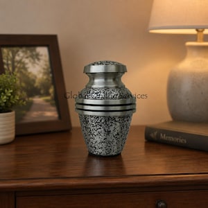 Op de afbeelding: Een zilveren en zwarte urn met een gespikkeld patroon. De urn heeft een deksel en twee horizontale banden in het midden. De urn staat op een houten oppervlak.