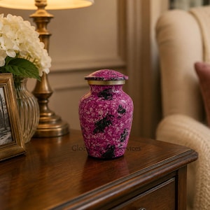 May include: A cremation urn with a vibrant magenta and white mottled finish, featuring black silhouettes of rabbits throughout. The urn has a metallic gold-toned rim at the neck and a matching lid with a rounded top, standing upright on a flat base.