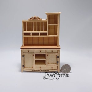 May include: A miniature wooden hutch with a light wood finish. The hutch has a top shelf with open compartments, a lower shelf with closed doors, and a bottom shelf with open compartments. The hutch is about 10 inches tall and is sitting on a white surface.