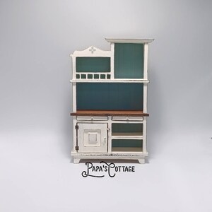 May include: A miniature white and teal hutch with a distressed finish. The hutch features a cabinet door, two drawers, and open shelving. The top section has a teal backing. The text "Papa's Cottage" is at the bottom.