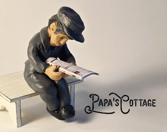 May include: A miniature figurine of a person in a dark outfit and cap, seated on a white bench, engrossed in reading a newspaper. The figurine is set against a plain white background. The text "Papa's Cottage" is visible.