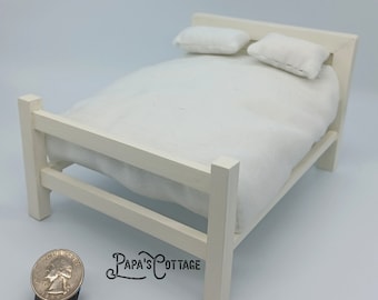 May include: White wooden dollhouse bed with a white mattress and two white pillows. The bed is about the size of a quarter.