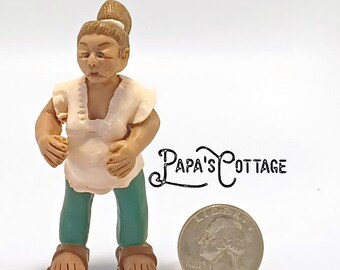 May include: A small, handcrafted figurine of a person wearing a cream-colored top, teal pants, and brown sandals. The figurine has a bun hairstyle. A quarter coin is next to the figurine. The text "Papa's Cottage" is visible.
