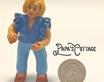 May include: A miniature figurine of a person wearing a blue top with ruffled sleeves and blue pants. The figurine has blonde hair and brown shoes. A quarter coin is next to the figurine. The text "Papa's Cottage" is also in the image.