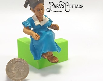 May include: A small figurine of a person in a blue dress with white trim, seated on a bright green block. The figurine has brown skin and hair. A quarter is next to the figurine. The text "Papa's Cottage" is in the background.