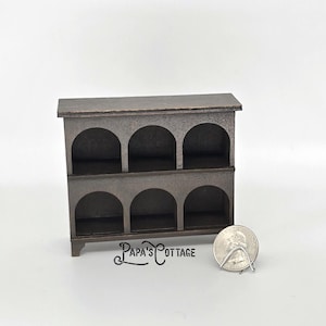 May include: A miniature wooden shelf unit with six arched openings, stained in a dark brown color. The text "Papa's Cottage" is visible on the front. A U.S. quarter coin is included for scale.