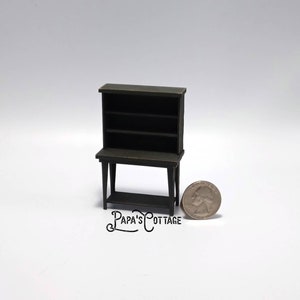 May include: Miniature dark green hutch with three shelves and a lower shelf, set on four legs. The hutch has a small top. A quarter is next to the hutch. The text "Papa's Cottage" is below the hutch.