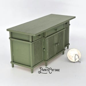 May include: A miniature sage green kitchen island with a countertop, drawers, and cabinet doors. The island has a textured wood grain appearance and is designed for a dollhouse or miniature setting. The text "Papa's Cottage" is visible.