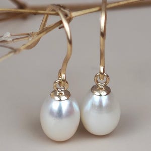 May include: A pair of gold-colored drop earrings with white, pear-shaped pearls. The earrings hang from delicate, curved hooks. The background is a soft, neutral color, with a sprig of dried flowers in the upper left corner.