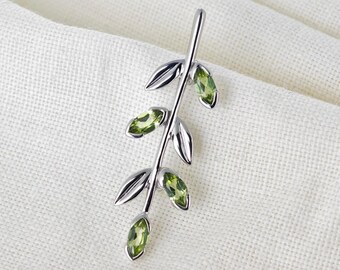 May include: A silver pendant shaped like a branch with five green gemstones. The pendant is on a white fabric background.