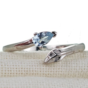 Può includere: Un anello d'argento con una pietra preziosa blu chiaro e un design a foglia. L'anello è su uno sfondo di tessuto bianco.