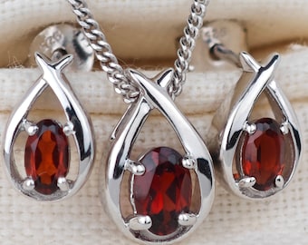 May include: A silver necklace and earrings set featuring a red gemstone pendant and matching earrings. The pendant and earrings are shaped like teardrops with a delicate silver design.