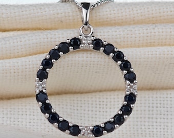 Può includere: Una collana con ciondolo in argento a forma di cerchio. Il cerchio è bordato da pietre preziose nere e ha un piccolo gioiello bianco in cima.