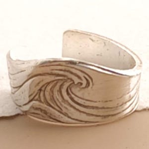 May include: A silver-toned open band ring featuring a textured, embossed design of a crashing ocean wave. The metal surface has a polished finish with recessed details that highlight the swirling motion of the water across the front of the ring.