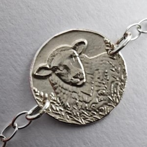 Op de afbeelding: Een zilverkleurige armband met een ronde bedel voorzien van een reliëf van een schapenkop. De bedel is verbonden met een fijne ketting van ronde schakels, wat zorgt voor een eenvoudig en samenhangend sieraad voor om de pols.