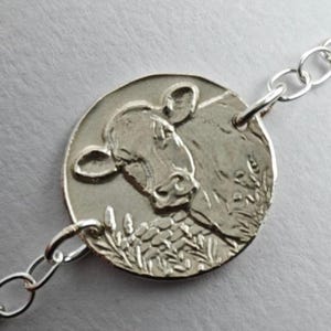 May include: A silver-toned bracelet featuring a circular charm with an embossed cow head design. The charm is centred on a delicate chain with round links. The metal has a polished finish, highlighting the detailed texture of the cow illustration.