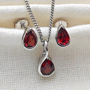 Puede incluir: Un conjunto de collar y pendientes de plata con piedras preciosas rojas en forma de lágrima. El collar tiene una cadena delicada y un colgante con un diseño retorcido.