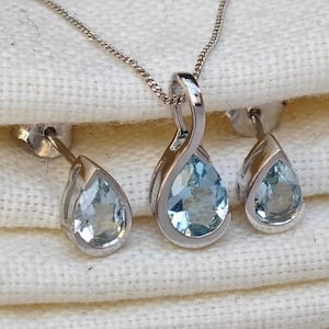 Op de afbeelding: Een sieradenset met een zilveren hangerketting en bijpassende oorbellen. De hanger en oorbellen hebben een druppelvorm met een lichtblauwe edelsteen. De set wordt gepresenteerd op een witte, getextureerde ondergrond.
