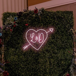 May include: A green wall backdrop with a black metal archway decorated with flowers. A pink neon sign in the shape of a heart with an arrow and the initials "L&D" is displayed on the wall.