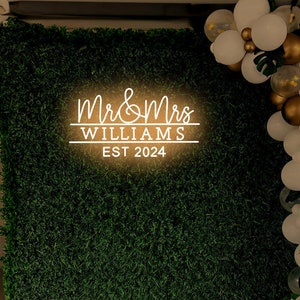 K&ouml;nnte beinhalten: Ein gr&uuml;ner k&uuml;nstlicher Gras-Hintergrund mit einem wei&szlig;en Neon-Schild, auf dem "Mr. & Mrs. Williams Est. 2024" steht. Rechts im Bild befindet sich eine Girlande aus wei&szlig;en und goldenen Luftballons.