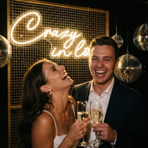 Op de afbeelding: Een lachend stel houdt champagneglazen vast. Achter hen een gouden raster met de neontekst "Crazy in Love". Hangende discoballen creëren een feestelijke sfeer.