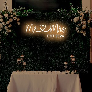 K&ouml;nnte beinhalten: Ein wei&szlig;es Neon-Schild mit den Worten "Mr & Mrs" und einem Herzsymbol, darunter der Text "EST 2024". Das Schild ist an einer gr&uuml;nen Wand mit Gr&uuml;npflanzen an beiden Seiten angebracht.