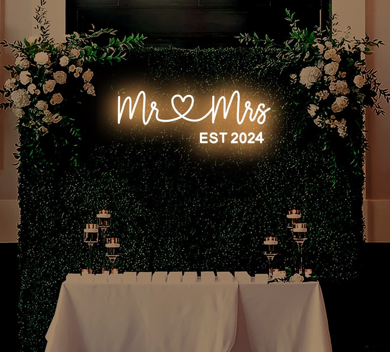 K&ouml;nnte beinhalten: Ein wei&szlig;es Neon-Schild mit den Worten "Mr & Mrs" und einem Herzsymbol, darunter der Text "EST 2024". Das Schild ist an einer gr&uuml;nen Wand mit Gr&uuml;npflanzen an beiden Seiten angebracht.