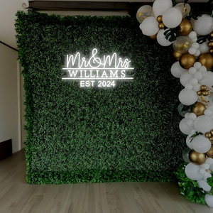K&ouml;nnte beinhalten: Ein wei&szlig;es Neon-Schild mit dem Text "Mr. & Mrs. WILLIAMS EST 2024" vor einem Hintergrund aus gr&uuml;nem Kunstgras. Wei&szlig;e und goldene Ballons sind rechts neben dem Schild zu einer Girlande angeordnet.