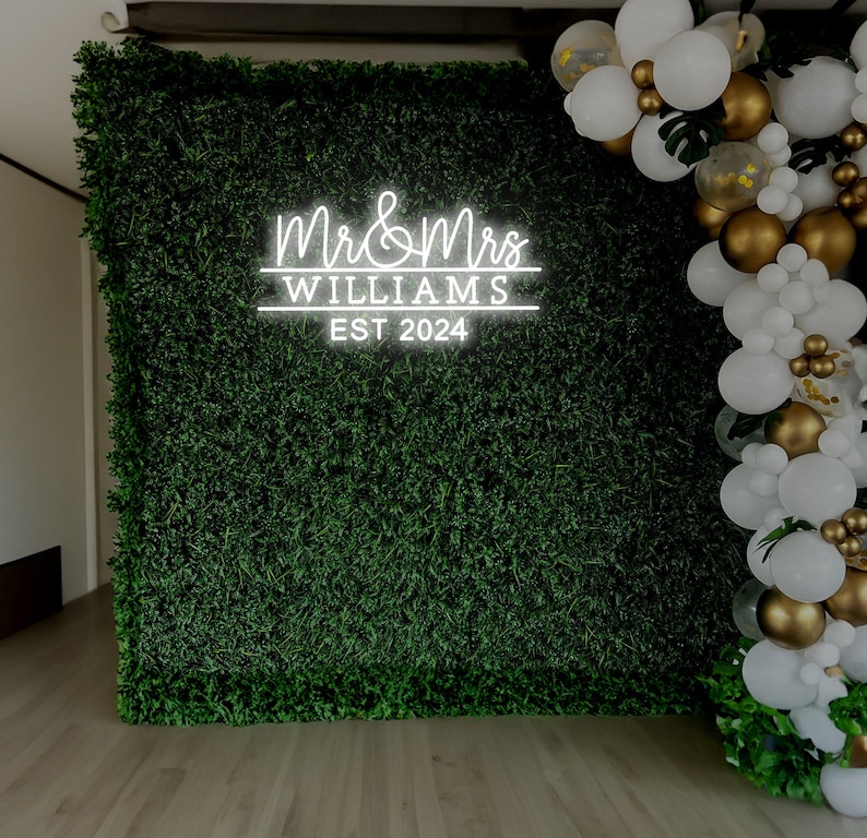 K&ouml;nnte beinhalten: Ein wei&szlig;es Neon-Schild mit dem Text "Mr. & Mrs. WILLIAMS EST 2024" vor einem Hintergrund aus gr&uuml;nem Kunstgras. Wei&szlig;e und goldene Ballons sind rechts neben dem Schild zu einer Girlande angeordnet.