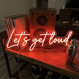 Op de afbeelding: Een rood neonbord met de tekst "Let's get loud" in een sierlijk lettertype. Een houten luidspreker, vinylplaten en een cocktailshaker zijn ook zichtbaar.