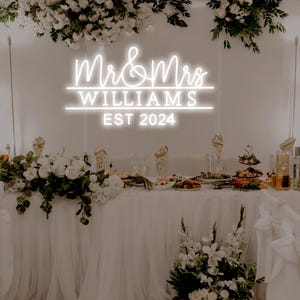 K&ouml;nnte beinhalten: Ein wei&szlig;es Neon-Schild mit dem Text "Mr. & Mrs. Williams Est. 2024" an einer wei&szlig;en Wand mit Blumendekoration. Das Schild befindet sich &uuml;ber einem wei&szlig;en Tisch mit wei&szlig;en Blumen und anderen Dekorationen.