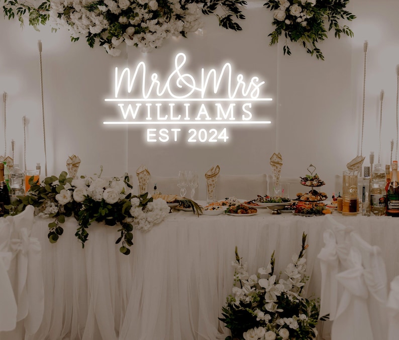 K&ouml;nnte beinhalten: Ein wei&szlig;es Neon-Schild mit dem Text "Mr. & Mrs. Williams Est. 2024" an einer wei&szlig;en Wand mit Blumendekoration. Das Schild befindet sich &uuml;ber einem wei&szlig;en Tisch mit wei&szlig;en Blumen und anderen Dekorationen.