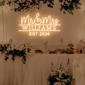 K&ouml;nnte beinhalten: Ein wei&szlig;es Neon-Schild mit dem Text "Mr & Mrs Williams Est 2024" an einer wei&szlig;en Wand. Das Schild wird mit einem warmwei&szlig;en Leuchten beleuchtet. Das Schild ist auf einem Tisch mit wei&szlig;en Tischdecken und Blumenarrangements aufgebaut.