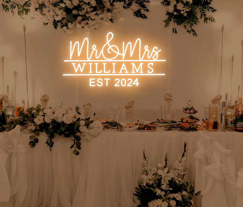 K&ouml;nnte beinhalten: Ein wei&szlig;es Neon-Schild mit dem Text "Mr & Mrs Williams Est 2024" an einer wei&szlig;en Wand. Das Schild wird mit einem warmwei&szlig;en Leuchten beleuchtet. Das Schild ist auf einem Tisch mit wei&szlig;en Tischdecken und Blumenarrangements aufgebaut.