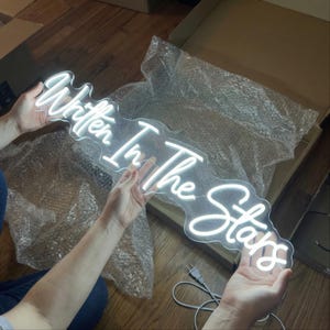 Op de afbeelding: Een wit neonbord met de tekst "Written In The Stars" in een cursief lettertype. Het bord wordt door twee handen vastgehouden, met een netsnoer zichtbaar. Het bord zit in een doos met bubbeltjesplastic.