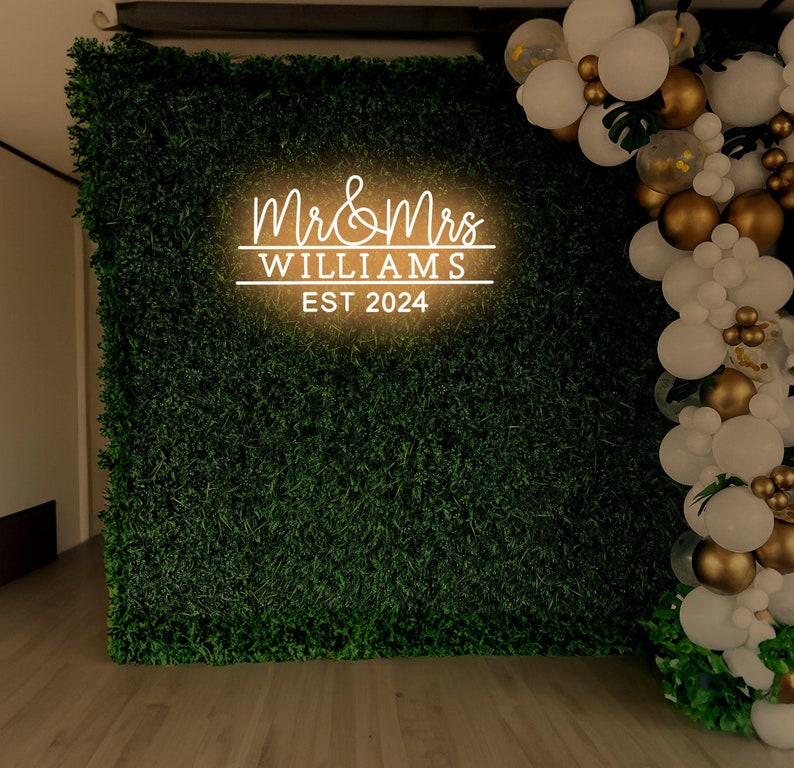 K&ouml;nnte beinhalten: Ein gr&uuml;ner k&uuml;nstlicher Gras-Hintergrund mit einem wei&szlig;en Neon-Schild, auf dem "Mr. & Mrs. Williams Est. 2024" steht. Rechts im Bild befindet sich eine Girlande aus wei&szlig;en und goldenen Luftballons.