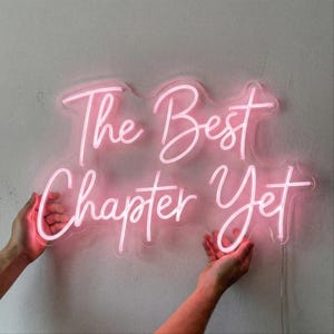 May include: A pink neon sign with the words "The Best Chapter Yet" in a cursive font. The sign is held up by two hands against a light gray background. The neon tubing is a bright pink color.