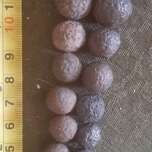 May include: A collection of 17 smooth, round, dark brown stones of varying sizes, arranged in a descending size order, with the largest stone at the top and the smallest at the bottom. A yellow measuring tape is visible on the left side of the image.