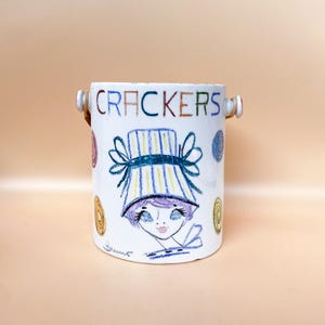 Puede incluir: Tarro de galletas de cerámica blanca con una ilustración dibujada a mano de una mujer con un gorro. La palabra "CRACKERS" está escrita en la parte superior. El tarro tiene dos asas pequeñas y está decorado con detalles coloridos.