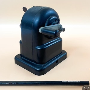 May include: A black vintage pencil sharpener with a metal crank and a small metal blade. The sharpener has a rectangular base and a round top.