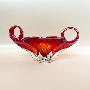 Op de afbeelding: Een rode glazen kom met twee handvatten. De kom heeft een heldere glazen basis en een rood en oranje swirl-ontwerp.