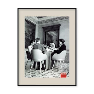 Könnte beinhalten: Schwarzweißfoto von vier Personen, die an einem Tisch in einem Raum mit hohen Decken sitzen. Die Personen sitzen auf weißen Stühlen mit schwarzen Beinen. Der Tisch ist mit einer weißen Tischdecke bedeckt. Der Raum hat einen großen Spiegel an der Wand und ein Gemälde an der Wand. Der Text "Cassina" ist unten im Bild rot.