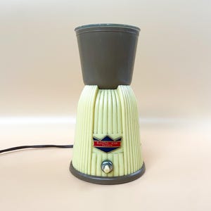 Può includere: Un frullatore per milkshake vintage Electro King con corpo in plastica bianca e marrone. Il frullatore ha un design a coste e un solo pulsante sul davanti. Il testo "Electro King Italiana" è stampato su un'etichetta blu e rossa.
