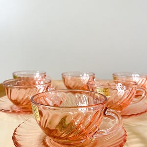 Op de afbeelding: Set van zes roze glazen theekopjes en schoteltjes met een swirl-ontwerp. De kopjes en schoteltjes zijn gemaakt van helder glas met een roze tint. De kopjes hebben een handvat en de schoteltjes hebben een gegolfde rand.