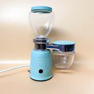 Può includere: Un macinacaffè elettrico vintage con finitura blu chiaro e cromata. Il macinacaffè ha un serbatoio per i chicchi di caffè in vetro, un contenitore in vetro per il caffè macinato e un timer.