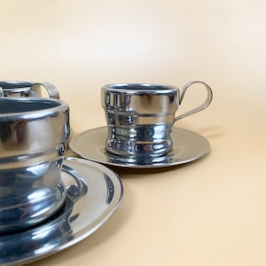 Peut inclure: Ensemble de trois tasses à espresso et soucoupes en métal argenté. Les tasses ont une forme arrondie et cylindrique et une petite poignée. Les soucoupes sont rondes et ont un bord légèrement surélevé.