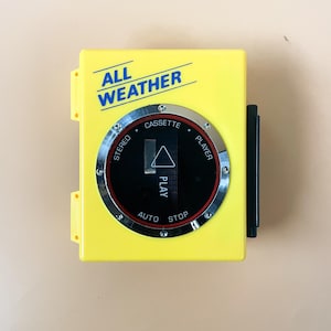 Peut inclure: Un lecteur de cassette jaune avec les mots "ALL WEATHER" imprimés sur le devant. Le lecteur a un logement de cassette noir et un bouton de commande argenté avec les mots "STEREO CASSETTE PLAYER", "PLAY", "AUTO" et "STOP" imprimés dessus.