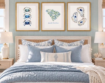 May include: A bedroom featuring a bed with a white quilted coverlet, a blue quilted blanket, and a cream-colored fringed throw. Three framed prints hang above the headboard: one with blue crabs, one with a floral South Carolina map, and one with oysters.
