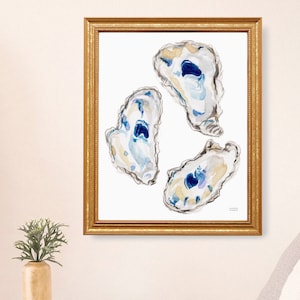 May include: A framed watercolor print featuring three oyster shells in shades of blue, beige, and white. The artwork is set against a white background and framed in a gold-colored frame. The art is displayed on a white wall.