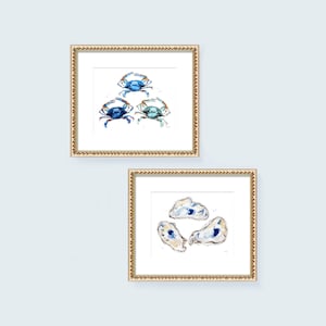 May include: Two framed watercolor paintings, one with blue crabs and the other with blue and white oysters. Both paintings have a gold frame.